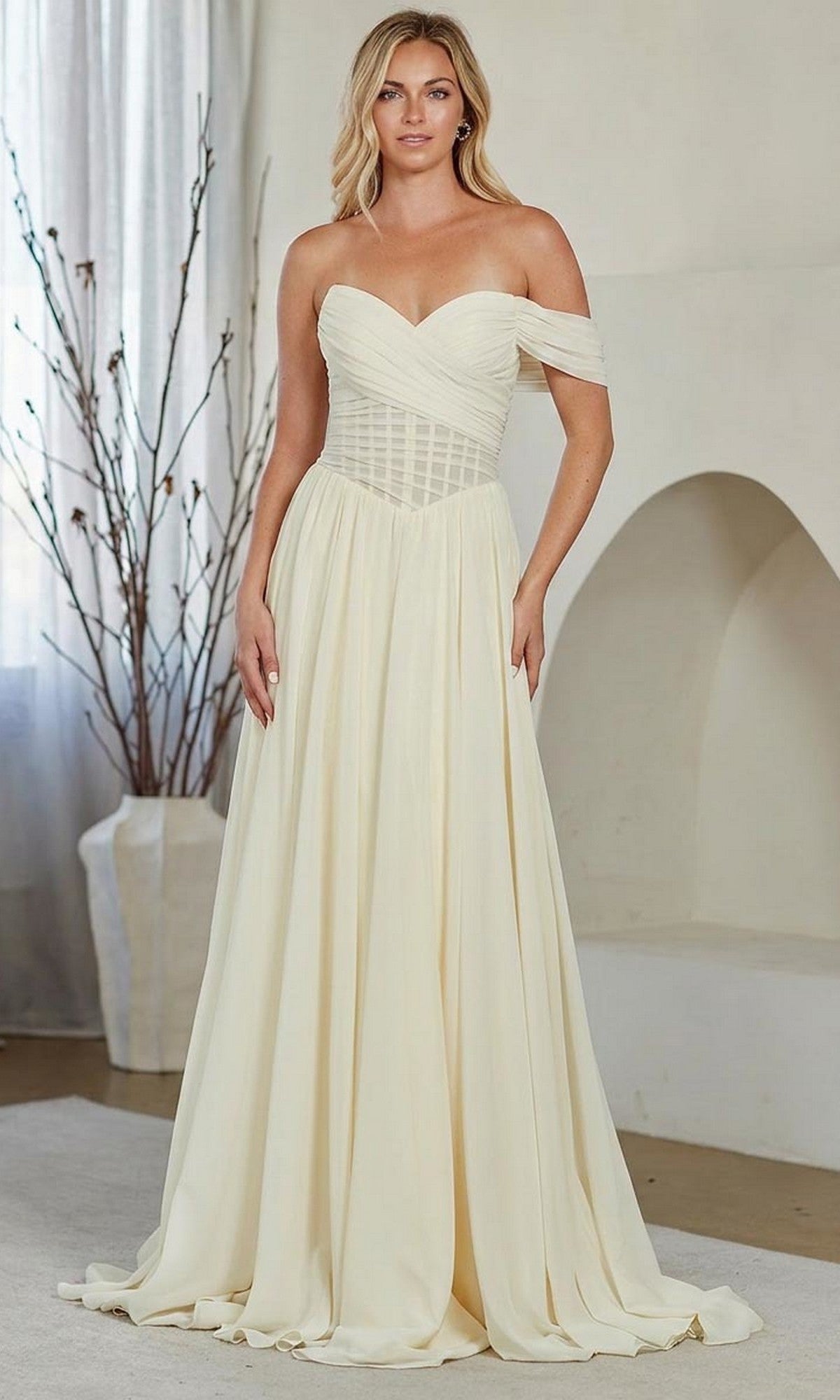 A woman stands indoors wearing the yellow Amelia Couture 9097 long prom dress, featuring an off-the-shoulder neckline, a fitted bodice, and a flowing A-line skirt.